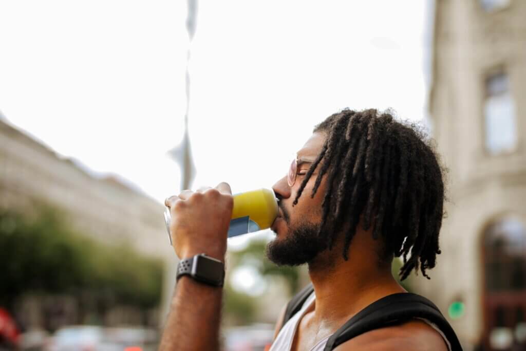 Black man with dreadlocks wearing a black smartwatch and sunglasses, drinking from a yellow can on a city street — representing smart tech in urban lifestyle.