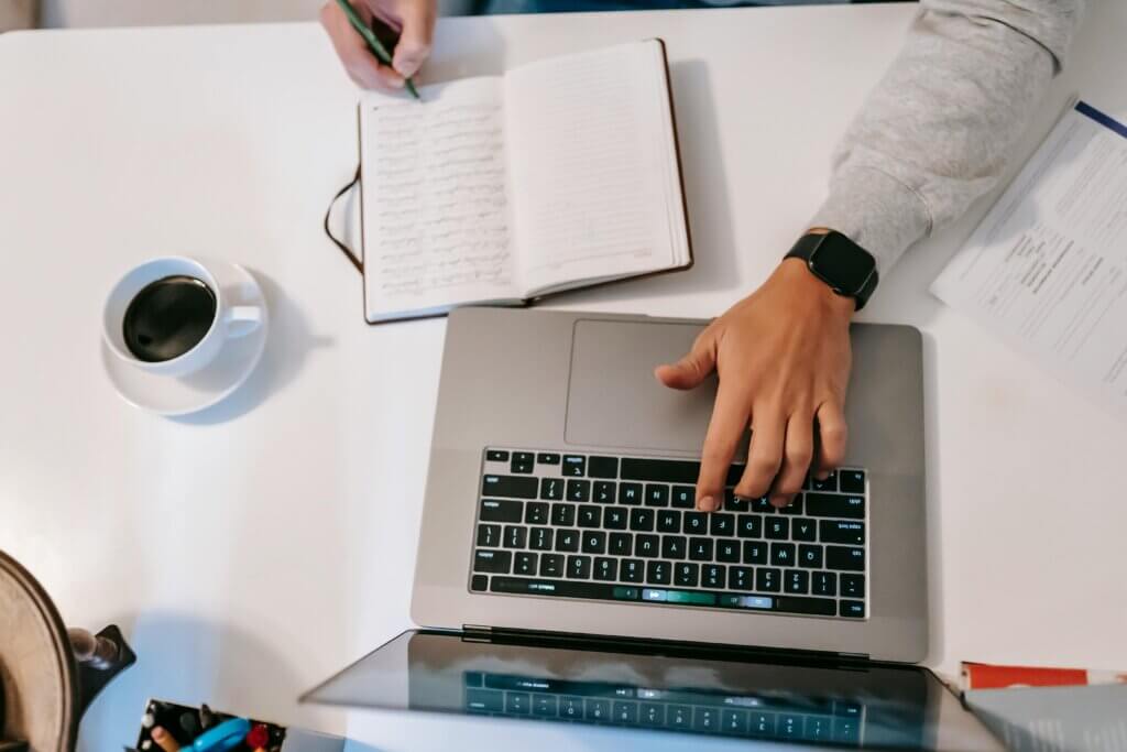 A man writing in a notebook while working on a laptop, wearing a black smartwatch, with a cup of black coffee on a white table.